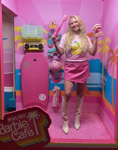 Woman posing in a bright pink Barbie-themed room holding a large toothbrush and dental model, representing a playful Barbie smile – barbie smile